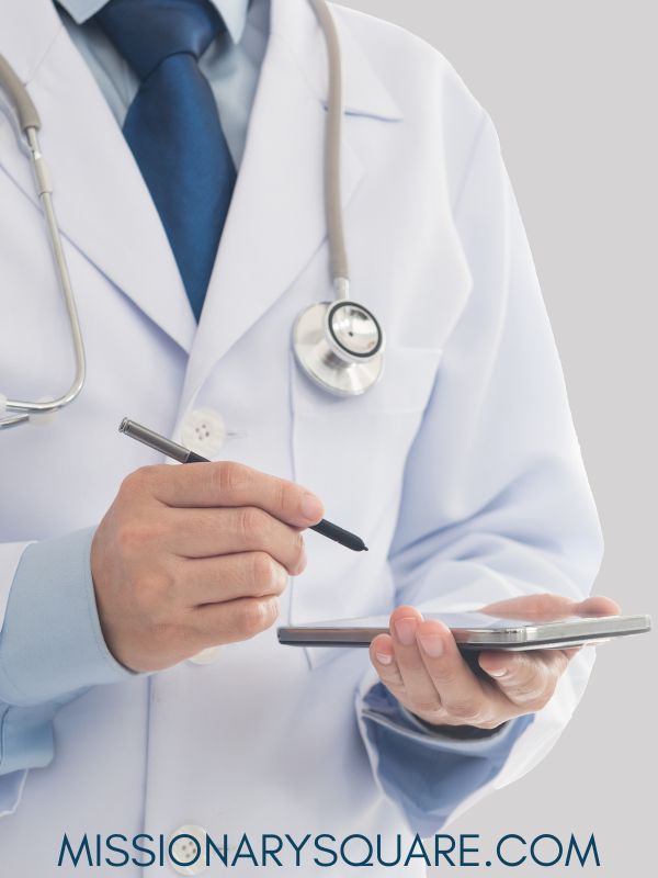 A closeup of a doctor from the shoulders down. He is wearing a stethoscope and writing with a pen on a tablet. He is wearing a blue tie. Text overlay says missinonarysquare.com 