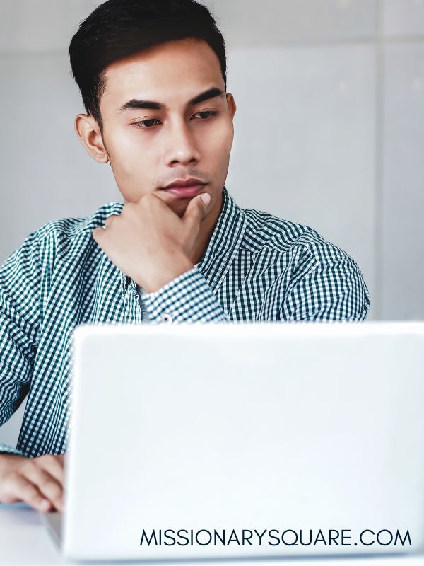 A young man looks at a laptop. He looks thoughtful and is touching his chin with his hand. He has dark hair and lighter skin. Text overlay says missionarysquare.com