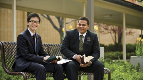 Two young men wearing suits and name tags for missionaries for The Church of Jesus Christ of Latter-day Saints sitting on a bench smiling off camera.